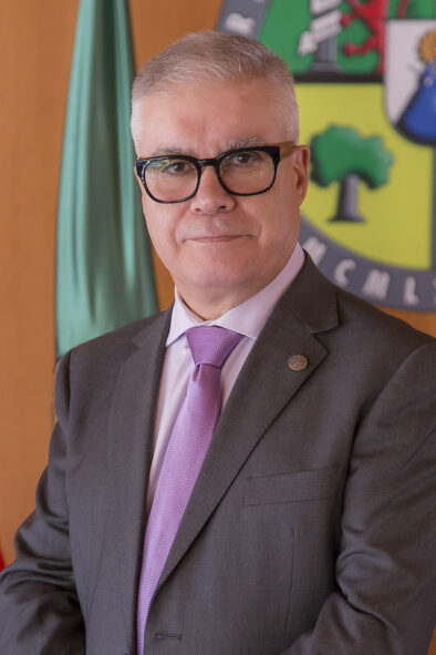 Foto retrato de Pedro Fernández Salguero con la bandera y el logo de la universidad detrás.